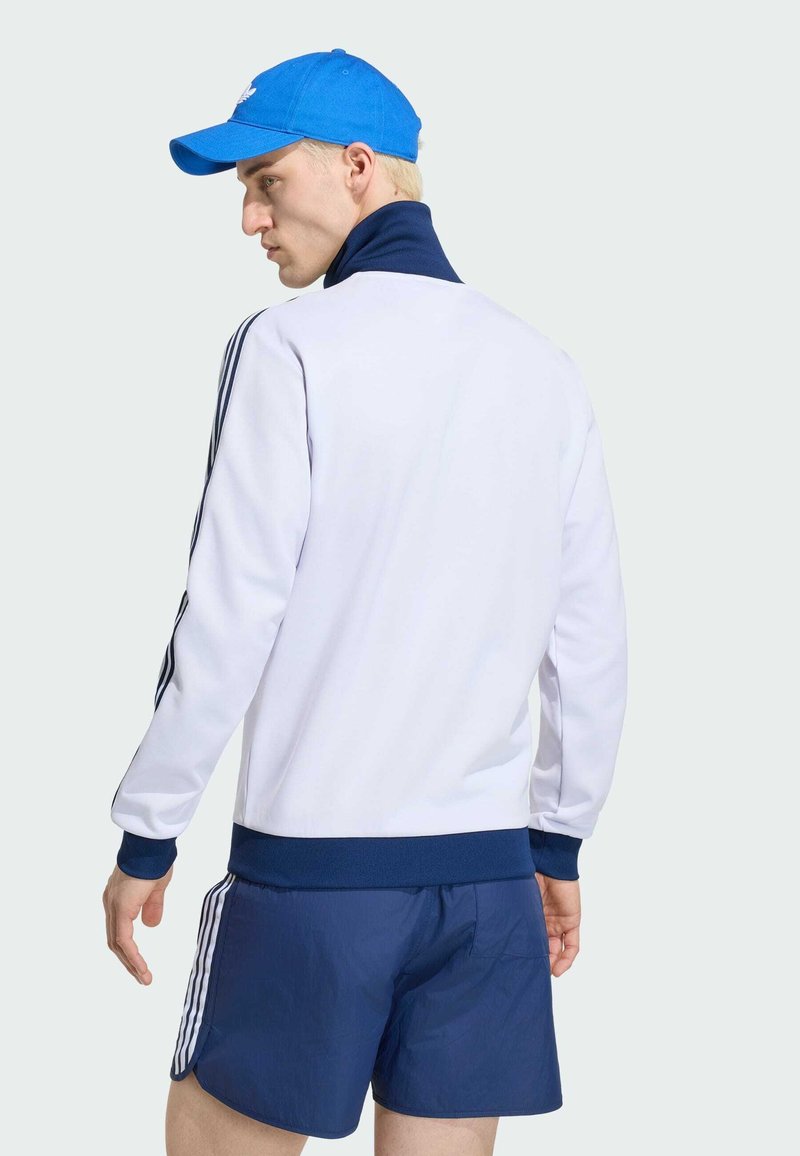 Hombre con chaqueta deportiva blanca con cuello y puños azul marino, pantalones cortos azul marino con franjas blancas y gorra azul, de perfil con la cabeza girada hacia atrás.