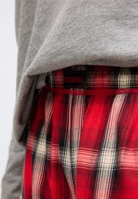 Sarkanas un melnas rūtainas pidžamas bikses ar baltām detaļām, ar elastīgu jostasvietu un melnu Diesel etiķeti. Pelēks audums izlīp pāri jostasvietai.