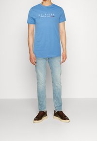 Muž v modrém tričku Hilfiger New York, světlých modrých džínách a tmavě hnědých teniskách stojící před jednobarevným bílým pozadím.