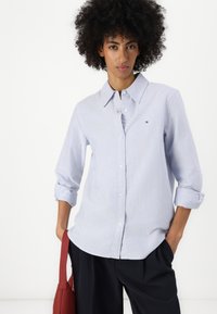 Modra in bela prugasta srajca s ovratnikom in zavihanimi rokavi, z gumbi in majhnim logotipom na prsih.