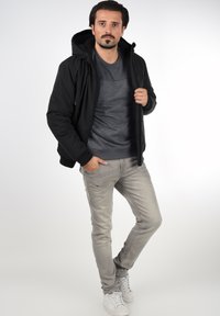 Veste à capuche noire avec une texture douce, superposée sur une chemise gris foncé avec un motif subtil ; associée à un jean gris clair et des baskets blanches.