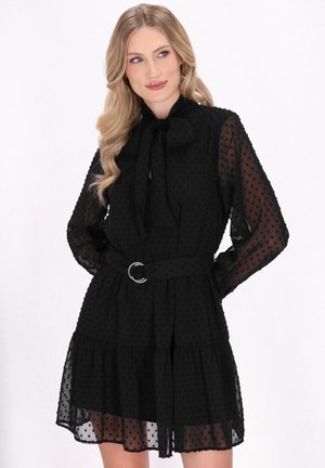 Rochie neagră din chiffon cu guler tip fundă, mâneci puff și tiv cu volane în straturi. Are o talie cu un centură prevăzută cu o cataramă din rhinestones și textură cu buline.