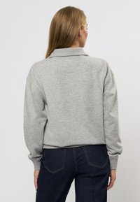 Svetrová mikina v šedej farbe s voľným strihom, so spustenými ramenami a rebrovanými manžetami. V kombinácii s tmavými denimovými džínami.