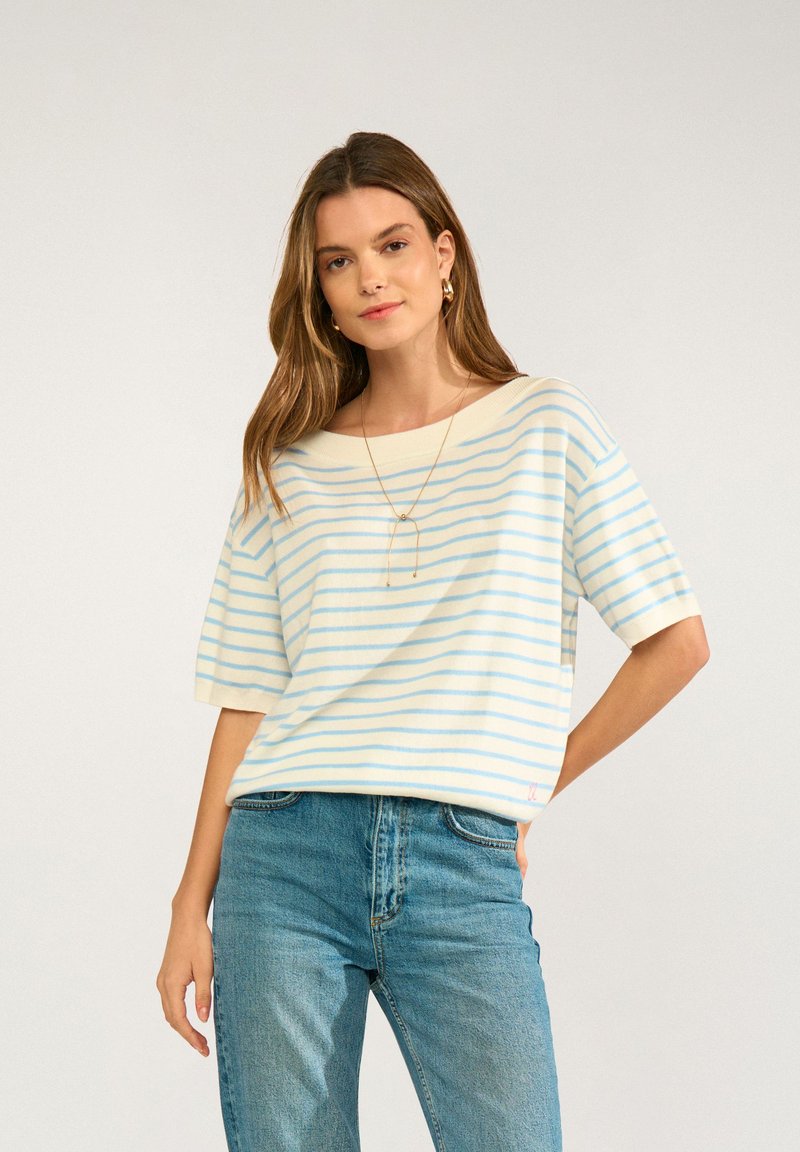 Jeune femme aux longs cheveux bruns portant un haut à manches courtes blanc et bleu clair rayé et un jean bleu, debout, une main sur la hanche.