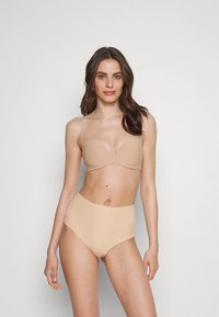 Beige spalvos vientisas liemenėlė su plonomis petnešėlėmis ir lygiu, lipdytu kaušelio dizainu, derinama su aukšta liemeniu beige spalvos apatiniais. Paprastas, minimalistinis stilius.