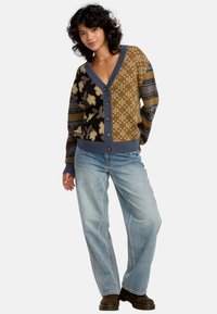 Strickjacke in Braun, Schwarz und Blau mit komplizierten floralen und geometrischen Mustern, V-Ausschnitt und Knopfverschluss. Kombiniert mit hellblauen Jeans.
