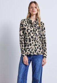 Beige Hemd mit Leopardenmuster und schwarzen Flecken, Knopfverschluss, lange Ärmel und Brusttasche, kombiniert mit blauen Jeans.