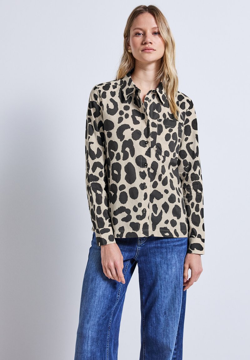 Beige Hemd mit Leopardenmuster und schwarzen Flecken, Knopfverschluss, lange Ärmel und Brusttasche, kombiniert mit blauen Jeans.