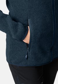 Marineblaue Fleecejacke mit gerippter Struktur, Reißverschlusstaschen und kontrastierenden dunklen Akzenten an den Bündchen und dem Saum. Nahaufnahme einer Hand in der Tasche.