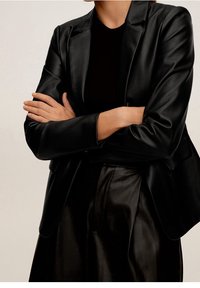 Blazer en cuir noir avec une coupe ajustée, des revers crantés et des détails de couture visibles, assorti à une jupe en cuir coordonnée.