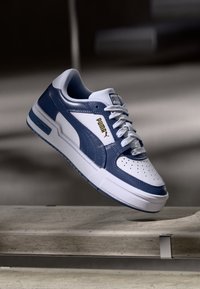 Blå og hvid lædersneaker med en perforeret tå, flade snørebånd og guld logo; har en tykk, hvid sål og slanke design detaljer.