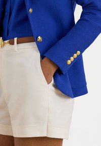 Lys blå teksturert blazer med gullknapper på ermene, kombinert med lyse beige shorts med sidelomme.