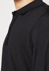 Polo noir à manches longues avec col et deux boutons noirs, tissu en maille texturée, porté par une personne visible du cou jusqu’au milieu du torse.