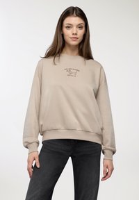 Beżowy sweter w luźnym kroju z ściągaczami, z haftowanym napisem w ciemniejszym brązie. Połączony z ciemnymi dżinsami.