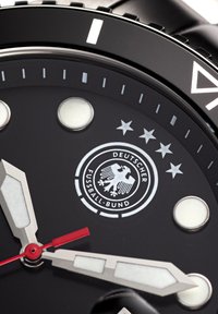 Schwarzes Zifferblatt mit silbernen und weißen Stundenmarkierungen, roter Sekundenzeiger und einem kreisförmigen Logo mit einem Adler und Sternen.