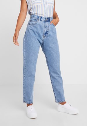 Person iført lyseblå højtaljede mom-jeans, hvide platform sneakers og en indstukket ærmeløs, stribet skjorte, der står med den ene hånd i lommen.
