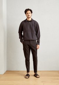 Svart zip-opp hettegenser med kengurulomme, kombinert med mørke cargo-bukser med sidelommer, og brune flate sandaler. Naturlig tregulv.