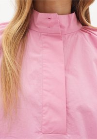 Kvinna som bär en rosa högkrageblus med långt blont hår som faller över ena axeln.