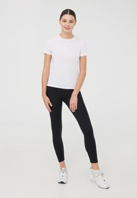Biała bawełniana koszulka zestawiona z czarnymi legginsami. Model nosi białe buty sportowe, prezentując swobodny, dopasowany styl z krótkimi rękawami.