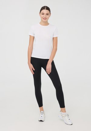 Biała bawełniana koszulka zestawiona z czarnymi legginsami. Model nosi białe buty sportowe, prezentując swobodny, dopasowany styl z krótkimi rękawami.