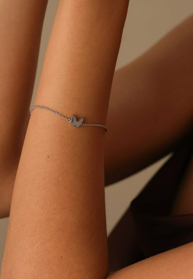 Braccialetto d'argento con un piccolo ciondolo a forma di farfalla, dotato di una catena delicata e superfici lisce e riflettenti, indossato su un braccio abbronzato.
