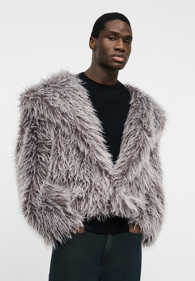 Jeune homme les mains dans les poches portant une veste en fausse fourrure grise à poils longs par-dessus une chemise noire, sur un fond blanc uni.