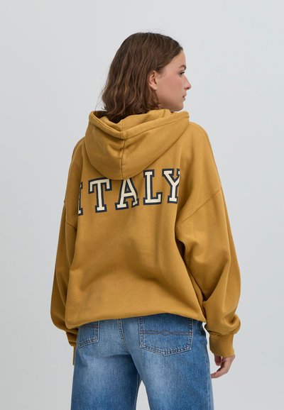 Dzeltena kapuce ar lielu baltu uzrakstu "ITALY" aizmugurē. Aprīkota ar aukliņu kapuces regulēšanai un brīvu piegriezumu. Savienota ar ziliem džinsu biksēm.