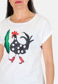Camiseta blanca con un pollo negro con lunares blancos, botas rojas y un diseño de hoja verde, cuello redondo y mangas cortas enrolladas.