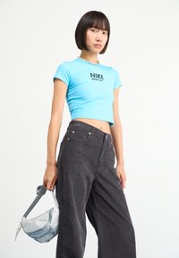 Light blue cropped Nike t-shirt with a black logo, paired with high-waisted dark gray corduroy pants. Model holds a gray crossbody bag.