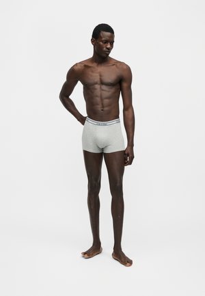 Homme torse nu debout pieds nus portant un boxer gris clair avec ceinture Calvin Klein sur fond blanc uni.