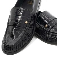 Zwarte loafers met een glanzende krokodillenstructuur, voorzien van een decoratief gouden embleem en franjes aan de voorkant. Soepele rubberen zool.
