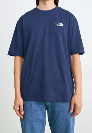 Personne portant un T-shirt bleu marine uni à manches courtes avec un petit logo blanc "The North Face" sur la poitrine gauche et un jean bleu.