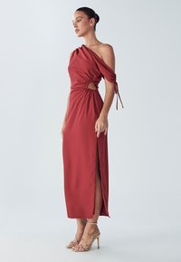 Sarkana plecu daļā nokarājusies kleita no gludas auduma, ar sānu šķēlumu un savākta dizaina detaļu ar atvērumu atslēgas formas, kopā ar zelta siksnu papēžiem.