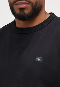 Svart sweatshirt i texturerad bomull, med rund halsringning och en liten logotyp-patch på framsidan nära kragen.