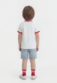 Παιδί με την πλάτη, φοράει λευκό πουκάμισο με κόκκινο περιθώριο, ανοιχτόχρωμα τζιν σορτς, κόκκινες κάλτσες με λευκές ρίγες και λευκά αθλητικά παπούτσια, μπροστά σε ομοιόμορφο φόντο.