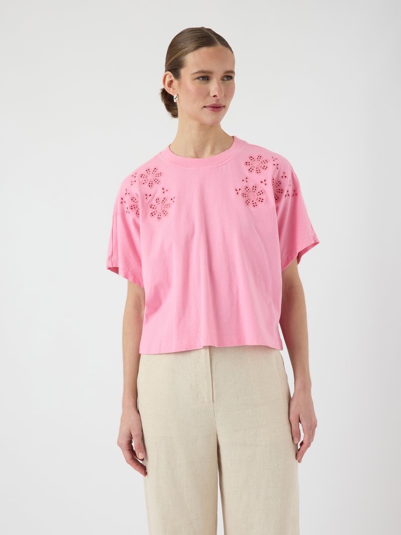 Tricou roz scurt cu modele florale decupate pe umeri, realizat din bumbac moale. Asortat cu pantaloni din in bej, având o croială relaxată.
