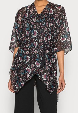 Frau trägt eine schwarze Wickelbluse mit Blumenmuster, dreiviertellangen Ärmeln und gebundenem Taillenband, kombiniert mit schwarzen Hosen.