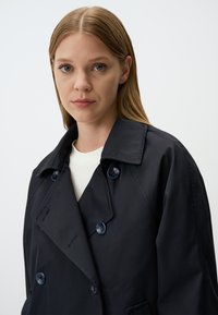 Granatowy dwurzędowy płaszcz trenczowy z dużymi ciemnoniebieskimi guzikami, klapą i gładką, matową teksturą. Prosta forma, minimalne zdobienia.