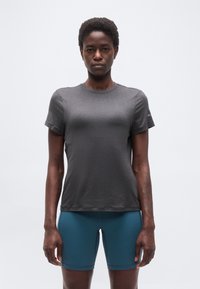 Άτομο με κοντά μαλλιά που φοράει εφαρμοστό σκουρόγκρι μπλουζάκι και τιρκουάζ σορτς ποδηλασίας, στέκεται με τα χέρια χαλαρά στο πλάι σε λευκό φόντο.