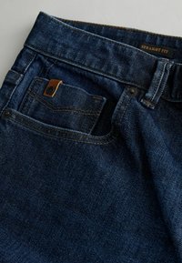 Proste dżinsy w ciemnoniebieskim kolorze, wyposażone w małą przednią kieszeń, zapięcie na miedziany guzik oraz kontrastowe przeszycia.