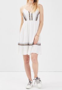 Weißes, ärmelloses Kleid mit gestickten vertikalen Streifen und einem Rüschensaum. Kombiniert mit weißen Turnschuhen für einen lässigen Look.