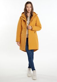 Žlutá zateplená parka s kapucí na stahování, zapínáním na zip a knoflíky, bočními kapsami a délkou ke kolenům. V kombinaci s modrými džíny a bílými teniskami.