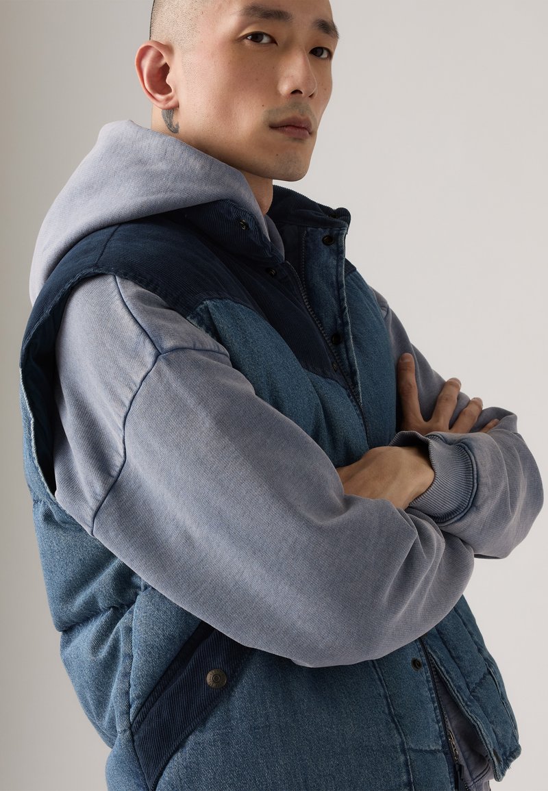 Denimvest med en grå hættetrøje; har et farveblokdesign i forskellige nuancer af blå, knaplukninger og ribbede manchetter.
