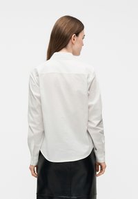 Γυναίκα με ίσια καστανά μαλλιά φοράει λευκό πουκάμισο με μακριά μανίκια και μαύρη φούστα, γυρισμένη προς τα πίσω, μπροστά σε απλό φόντο.