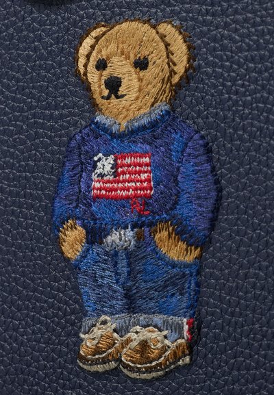 Mackófigura hímzés kék pulóverben, amelyen amerikai zászló látható, kék farmerben és barna tornacipőben, sötétkék, texturált anyagon.