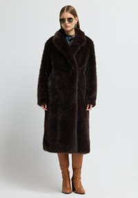 Abrigo de piel sintética marrón con cuello de chal, frente de doble botones y mangas largas. Lleva puesto unas botas de tacón color beige hasta la rodilla.