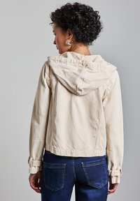 Beige tyyppi lyhyt takki hupulla, jossa on napilla varustetut hihansuut ja sivutaskut. Puettu tummansiniisiin farkkuihin. Sileä rakenne.