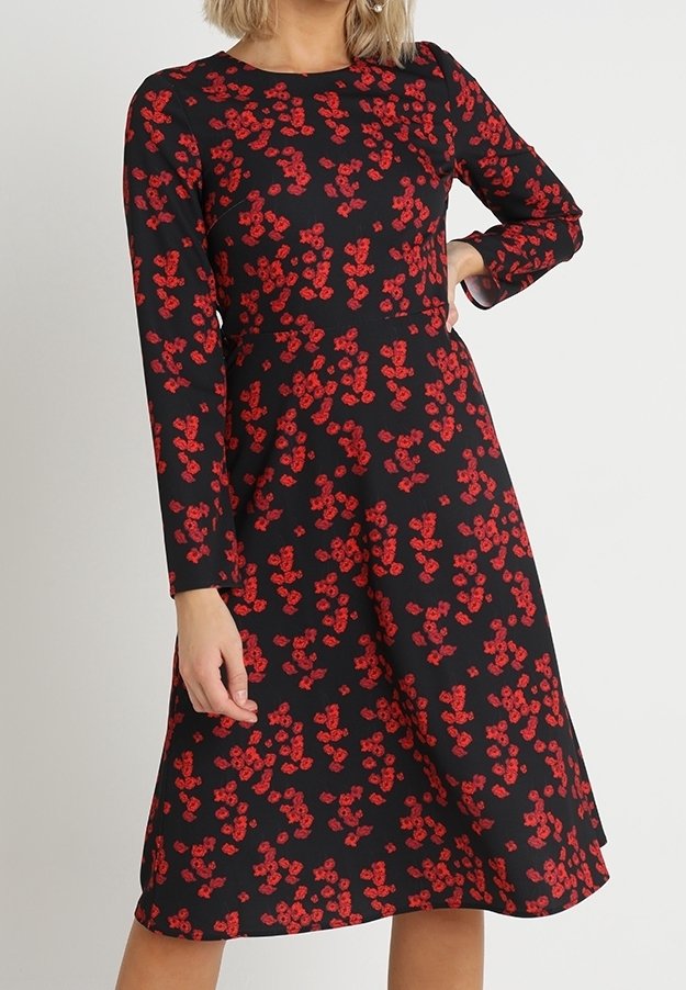 Robe midi noire à manches longues avec motif floral rouge, taille ajustée et jupe trapèze, portée par une personne avec une main sur la hanche.