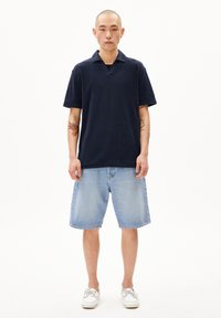 Marineblauwe katoenen polo-shirt met korte mouwen en open kraag, gecombineerd met lichtblauwe denimshorts en witte bootschoenen.