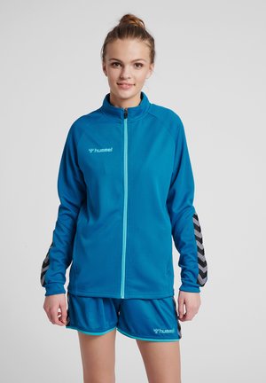 Ung kvinde iført blå sportjakke og matchende shorts, står og vender fremad mod en ensfarvet grå baggrund.